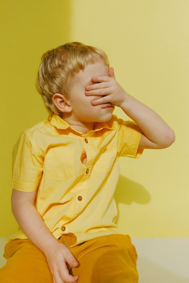 Een kind in een geel blousje met zijn linkerhand hand voor zijn ogen en neus. In een gele kamer